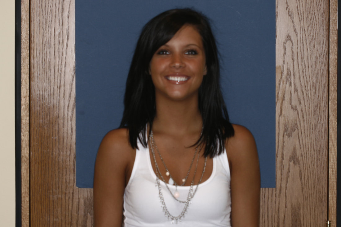 Smiling woman wearing white tank top and silver necklace against blue background with wood framing.