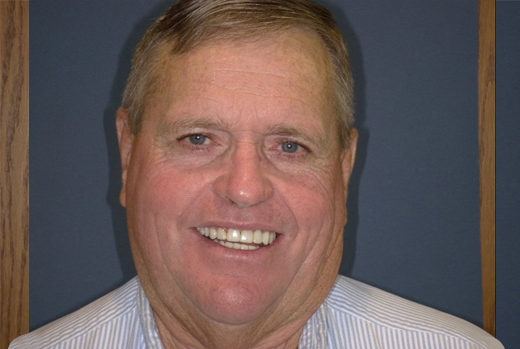 Middle-aged man smiling at camera in professional portrait against blue background wearing light blue striped shirt.