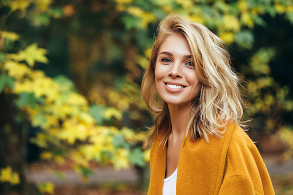 Woman smiling outdoors wearing yellow cardigan with blurred autumn foliage background.