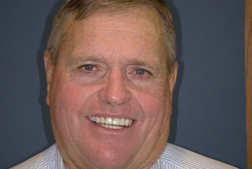 Smiling man in light blue shirt against gray background, professional headshot for dental practice.