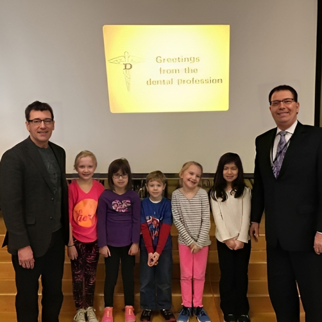 Two dental professionals posing with five children in front of projection screen displaying dental profession greeting.