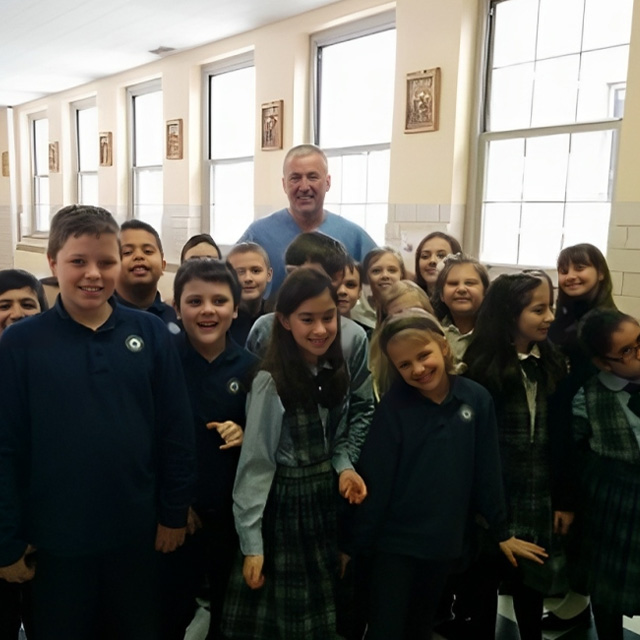 Adult man with group of smiling children in school classroom with large windows.