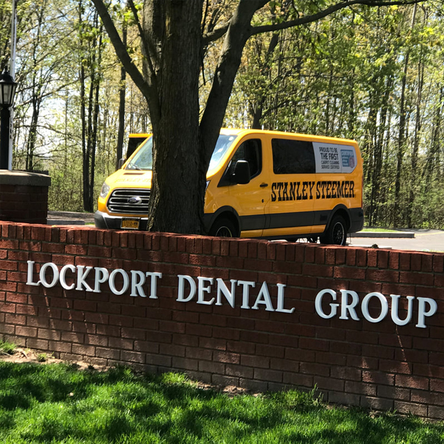 Yellow Stanley Steemer van parked at Lockport Dental Group brick sign entrance with trees.