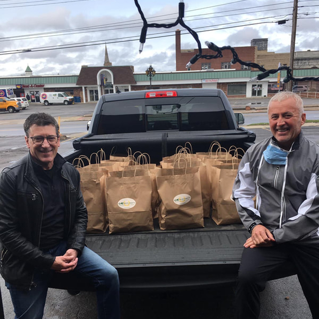 Two men pose in pickup truck bed filled with brown paper bags at community donation event.