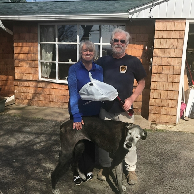 Two people posing with a dog outside a brick cottage, holding a white bag.