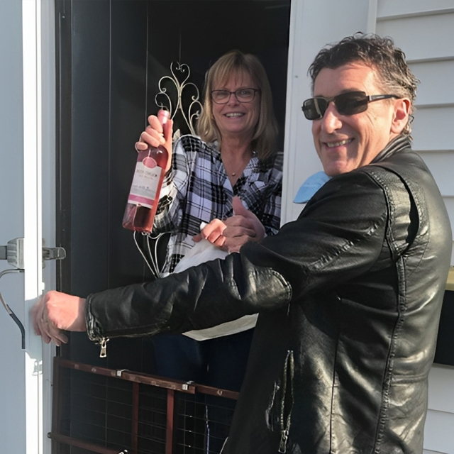 Two people holding a bottle of rosé wine at a doorway, smiling at the camera.