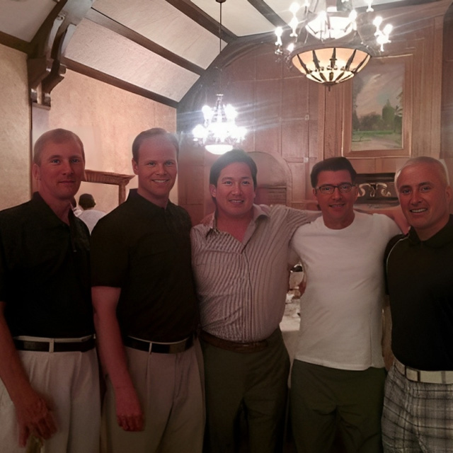 Five men posing together in an elegant interior room with wood beams and chandelier lighting.