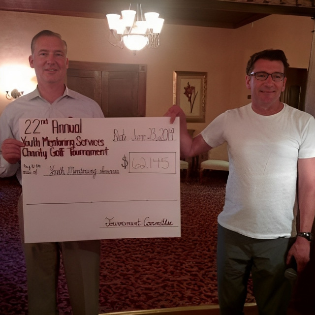 Two men holding oversized check for $20,145 from 22nd Annual Youth Mentoring Services Charity Golf Tournament indoors.