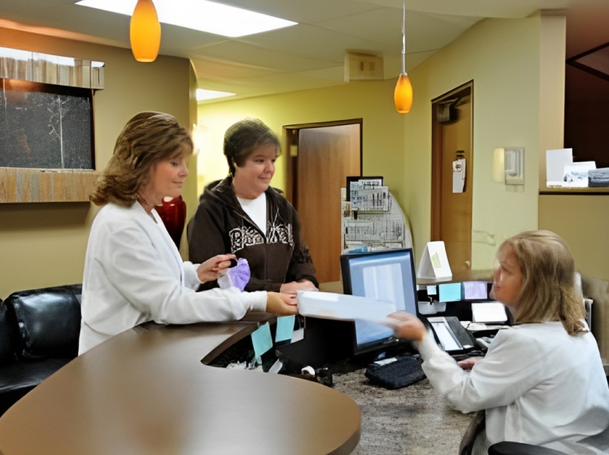Three women in dental office reception area exchanging documents at desk.