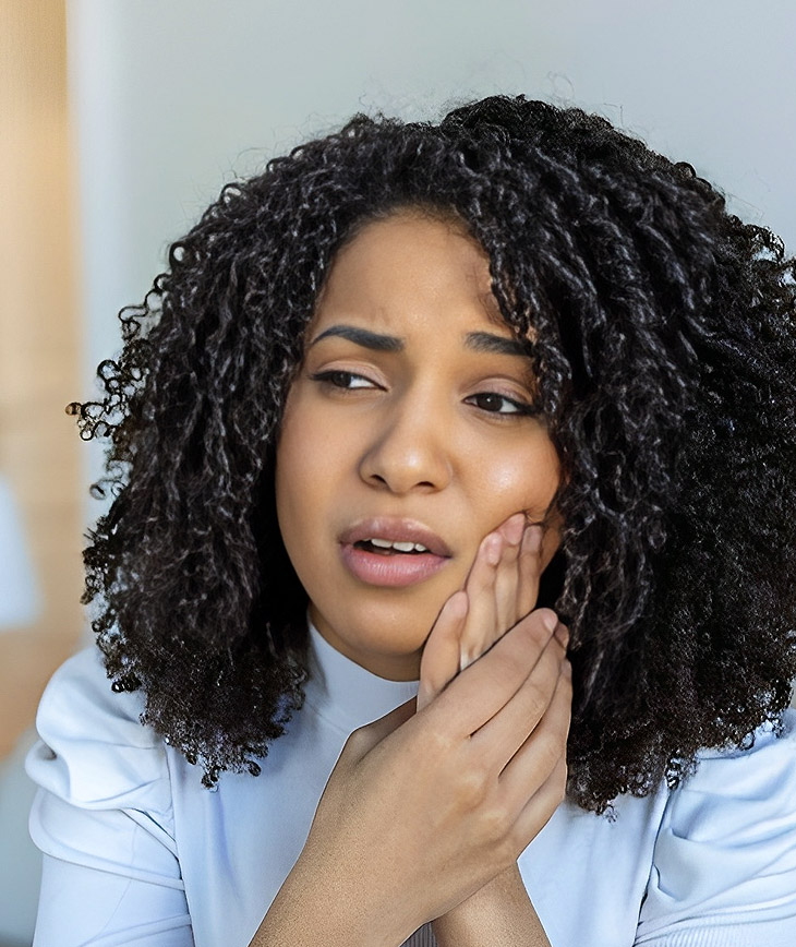 Woman with curly dark hair touching her cheek, appearing to have tooth pain or discomfort.