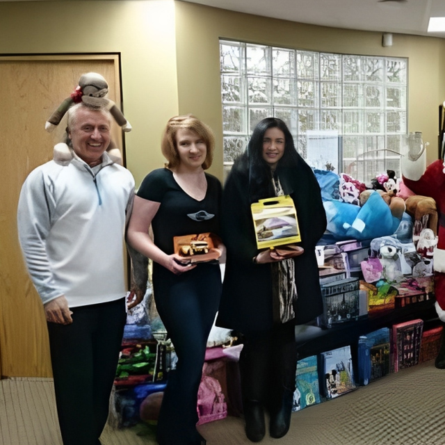 Three people posing with donated toys and gifts in an indoor office setting.
