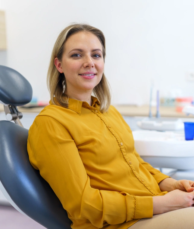 Professional woman in yellow shirt seated in dental office chair, smiling at camera.