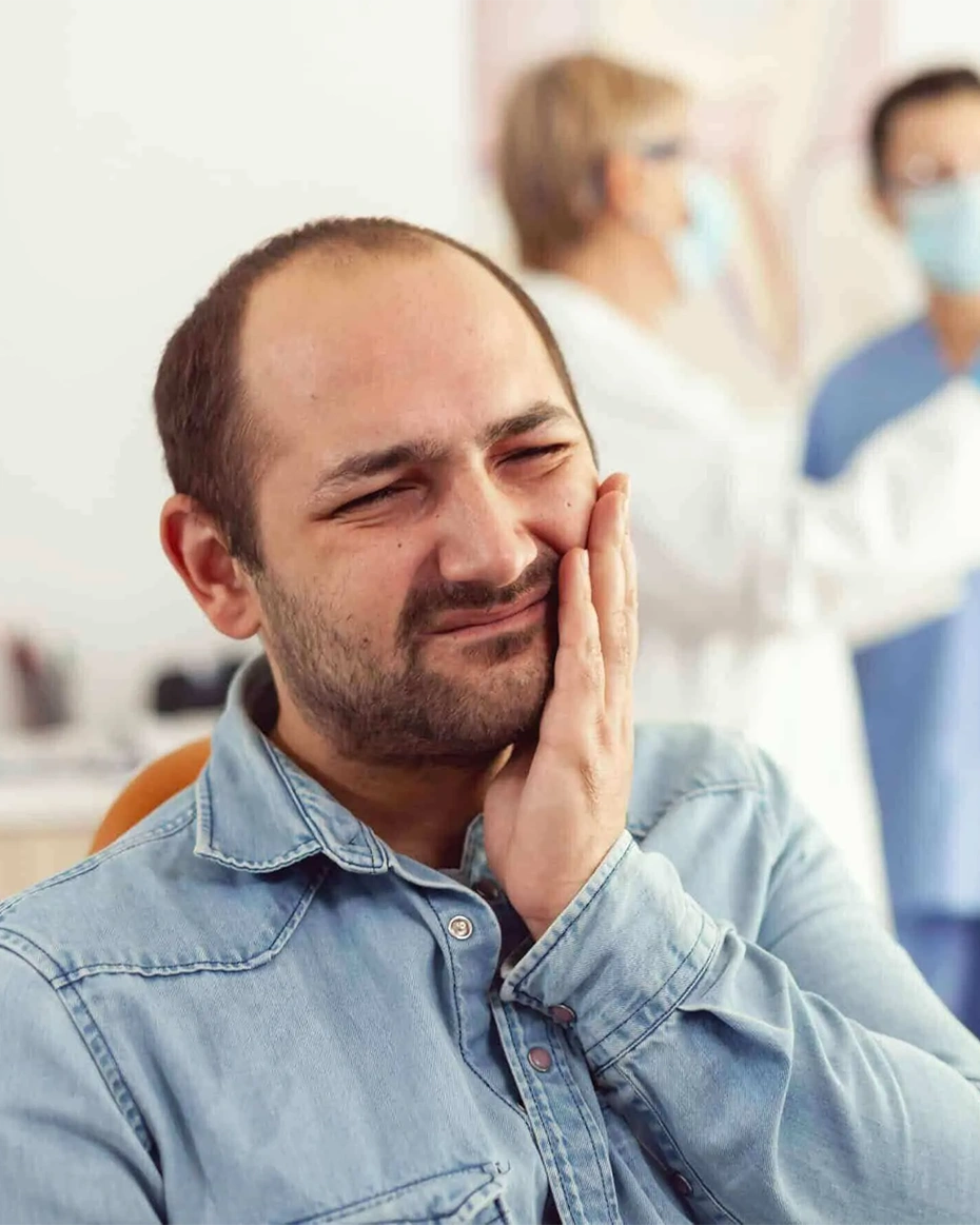 Man experiencing tooth pain in dental office with healthcare professionals in background.